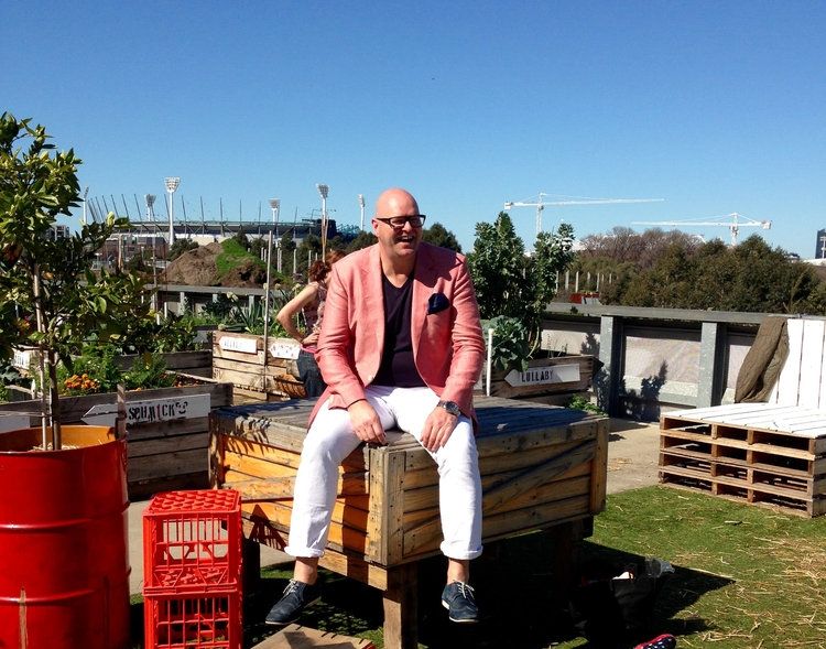 Shot of man sitting on the corner of a wooden crate wearing a pink jacket over a black top and white pants.. Looks to be on a rooftop terrace with the MCG in the background.
