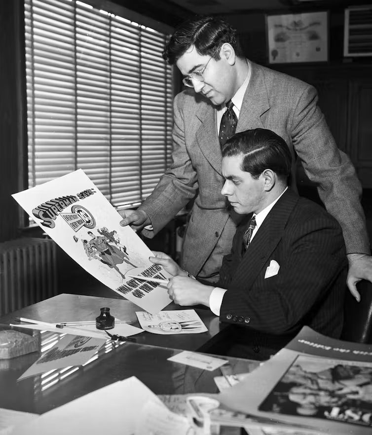 Jerry Siegel and Joe Shuster - the creators of the Superman character, are both looking at a poster with the comic drawing of Superman and a large title at the top reading "SUPERMAN SAYS".