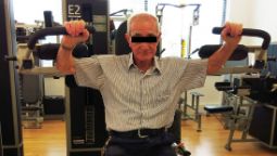 An elderly male is doing lat raises on a piece of exercise equipment in a gym