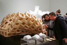 Two people study a large wooden wave, made from interlocking cubes. Small plastic versions of the structure in different wave forms sit on the table in front.