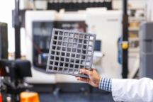 A man holds a square crosshatched 3D printed grey material in front of a 3D printer in a lab