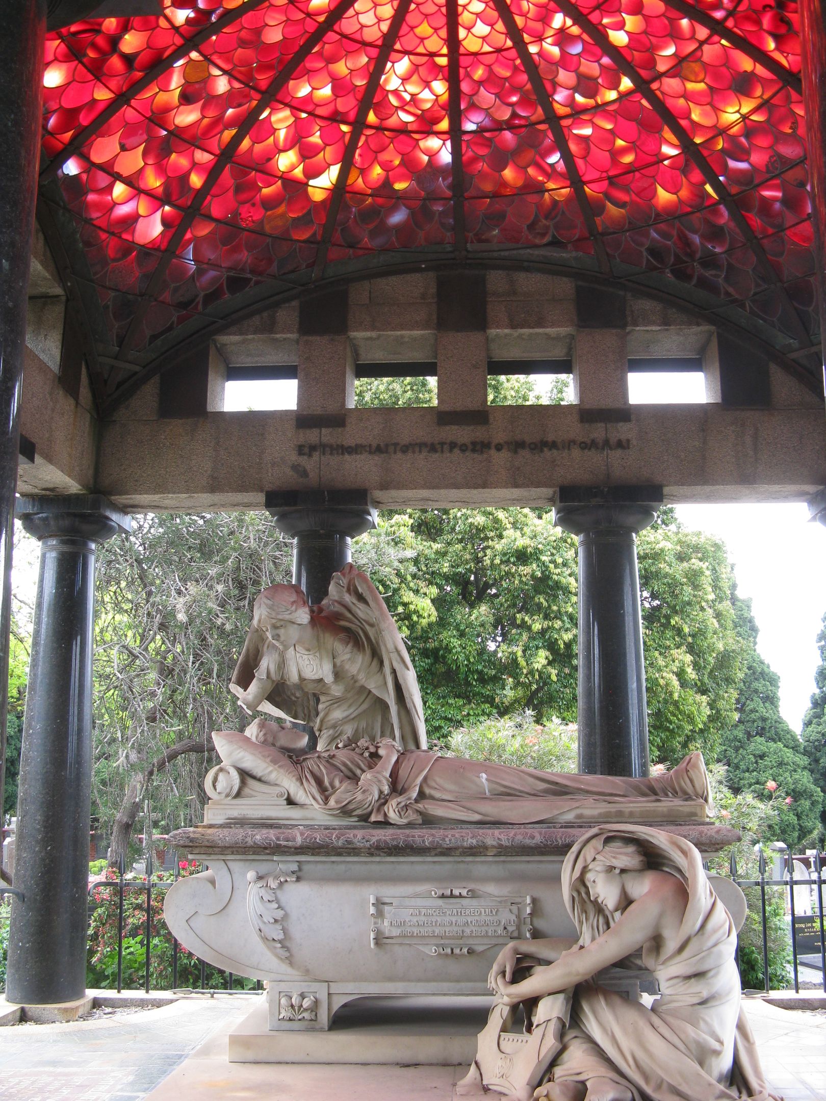 A statue consisting of a man lying down on a bed, with an angel leaning over him and another woman crouched at the foot of the bed. All are dressed in ancient civilisation style clothes. The status is inside a rotunda in a park with red stained glass on the roof. 