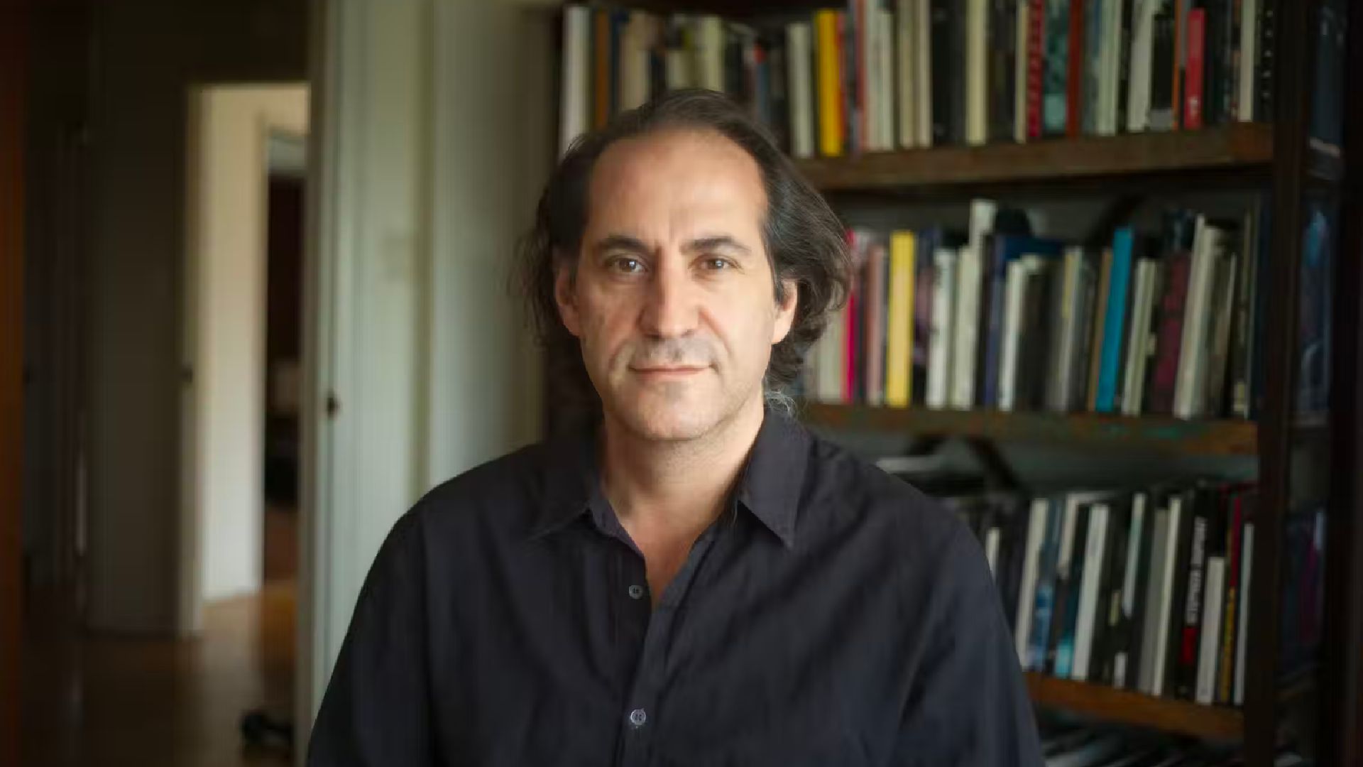 A photo from the chest up of Author Steve Toltz, he has a calm, neutral expression and long dark hair pushed away from his face