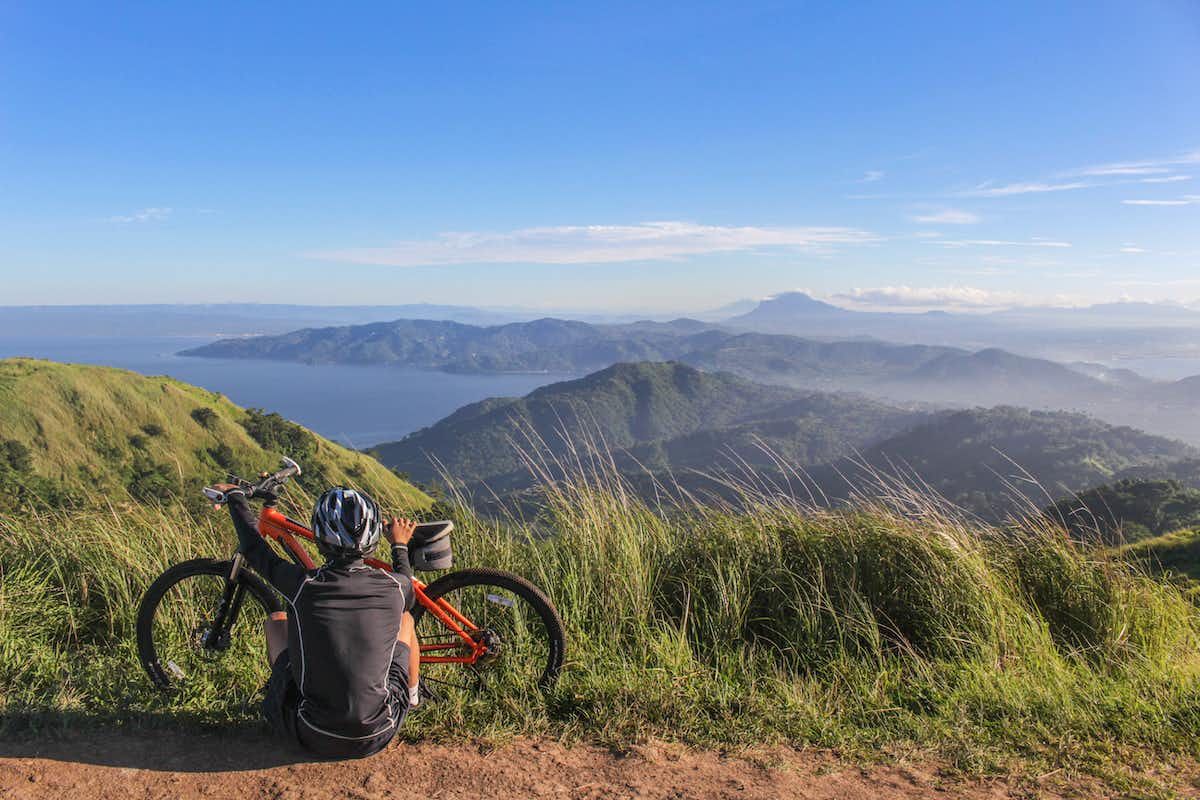 A bike rider sitting on the ground next to a bike on top of a hill looking out towards the view of mountains and the sea.