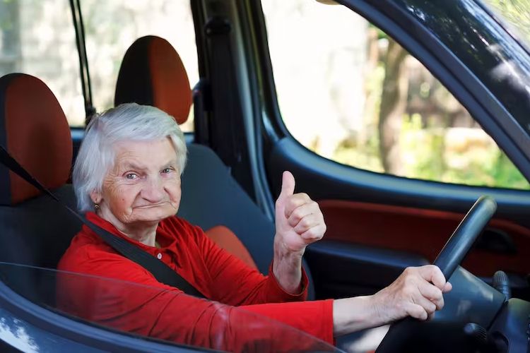 Elderly woman driving a car, looking at the camera smiling and holding her left up, giving the thumbs up