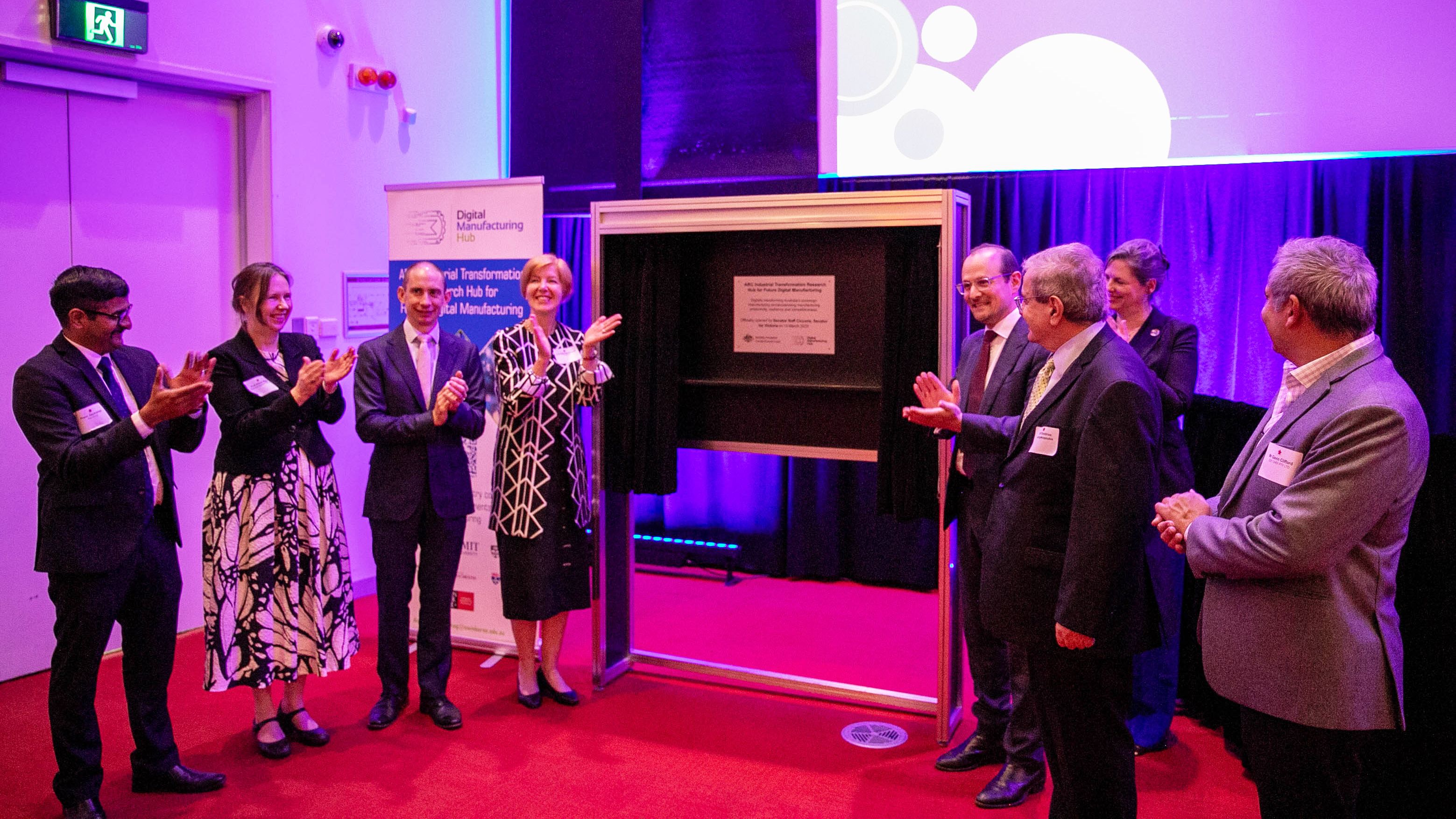 Eight people stand at a launch event clapping hands next to an official plaque.