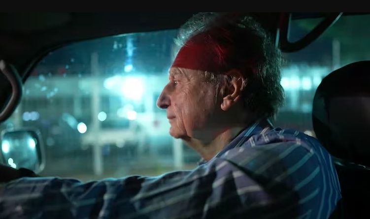 Side profile of an elderly man driving a car at night, with white hair and a blue striped shirt