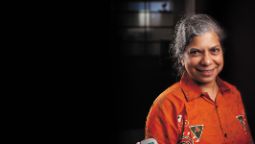 A female with greying hair is wearing an orange shirt and stands against a dark background.