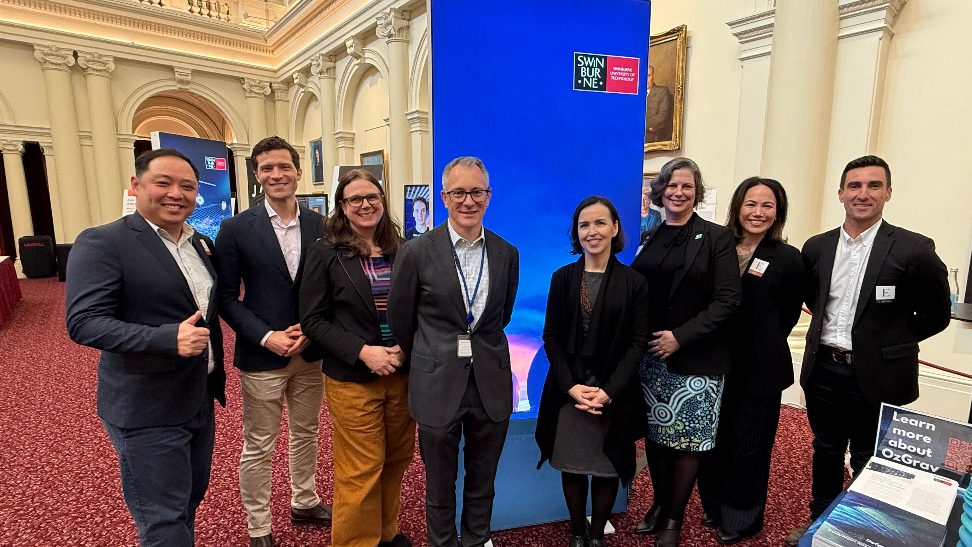 8 professional people in front of a Swinburne banner in an official parliament building.
