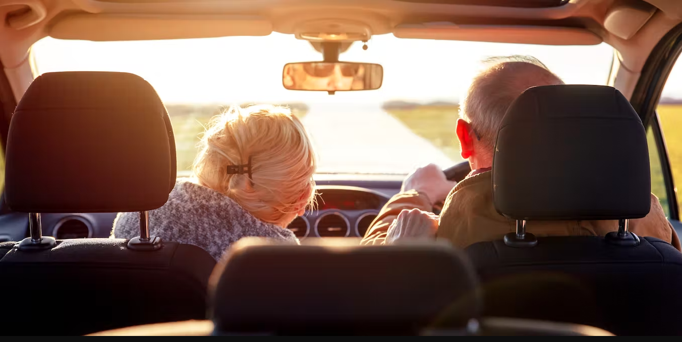 An elderly couple in a car, the man is driving on the right and the women is in the front passenger seat to the left. The sun is glarly through the front car window. Shot is from the back middle passenger seat.