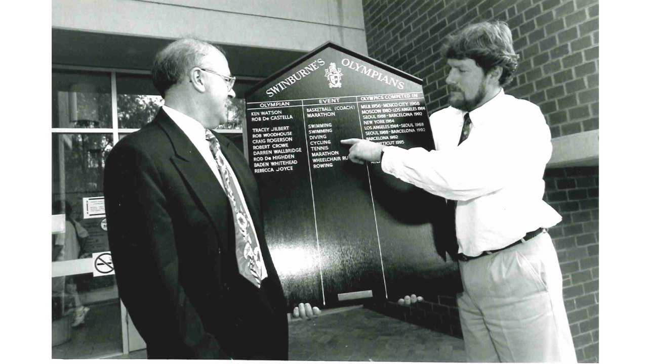 Bruce McDonald standing in front of the honour roll of Swinburne Olympians to Sports Association Executive Officer with Andrew Tingate that he donated in 1996. 