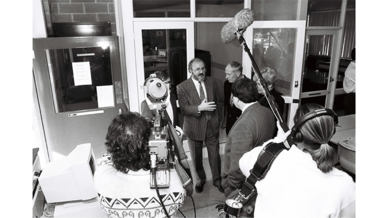 A black and white image of Bruce McDonald standing in front of a camera crew at the Wantirna Campus celebration for Proclamation Day, 1992  