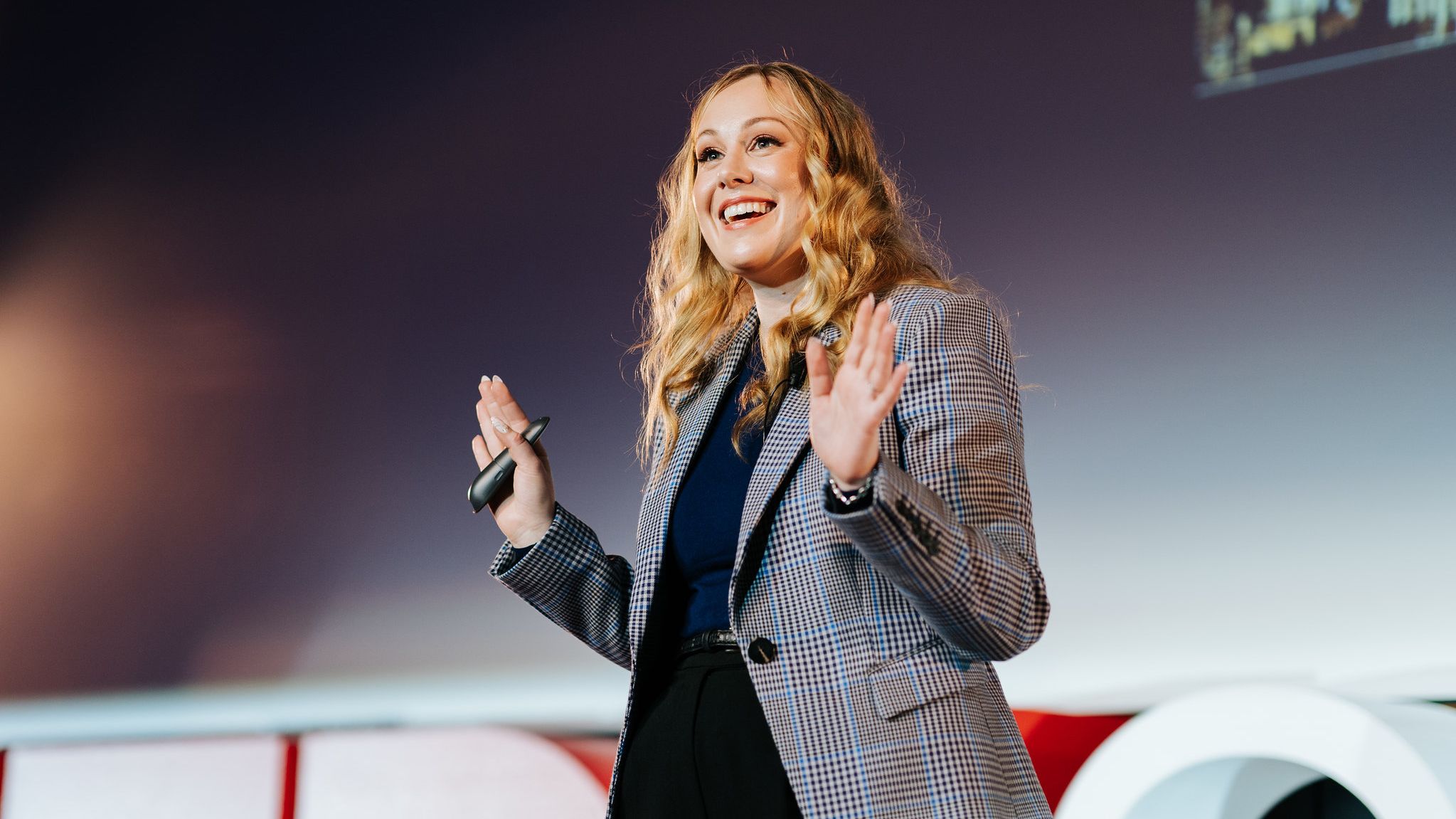 A person stands on stage with hands raised, one containing a presentation remote.