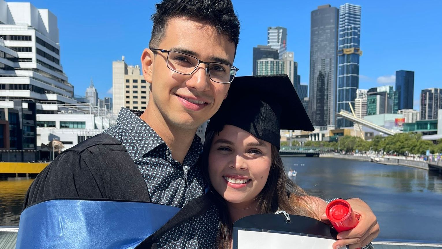 A man and woman are in front of the Melbourne skyline they are wearing graduation gowns