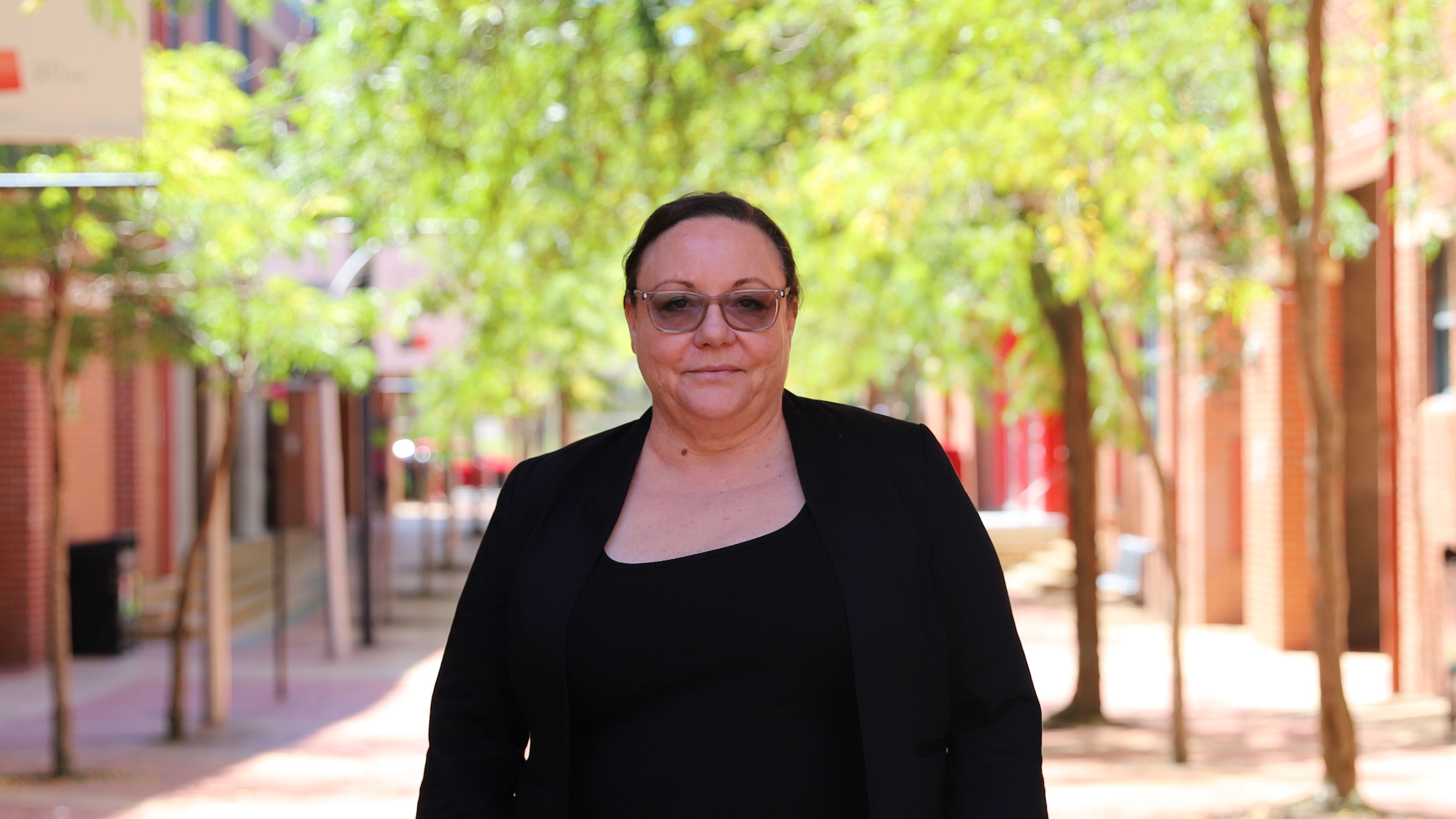 Director, Indigenous Engagement, Vicky Peters is pictured from the waist up. Her dark clothing contrasts against a bright, tree filled background on Swinburne's hawthorn campus