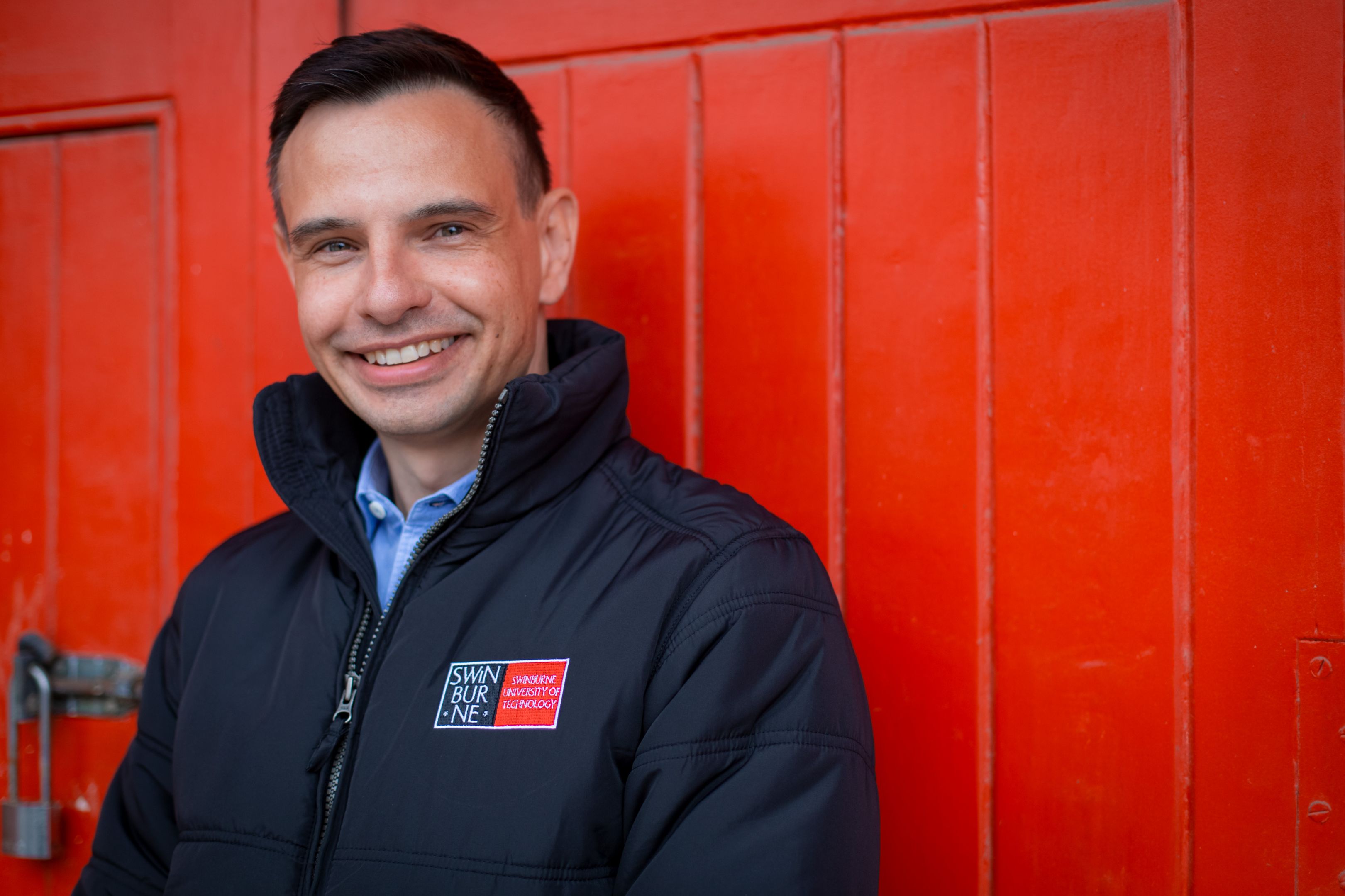 Werner smiles to camera wearing a black jacket featuring the SUT logo with a bright red wall in the background