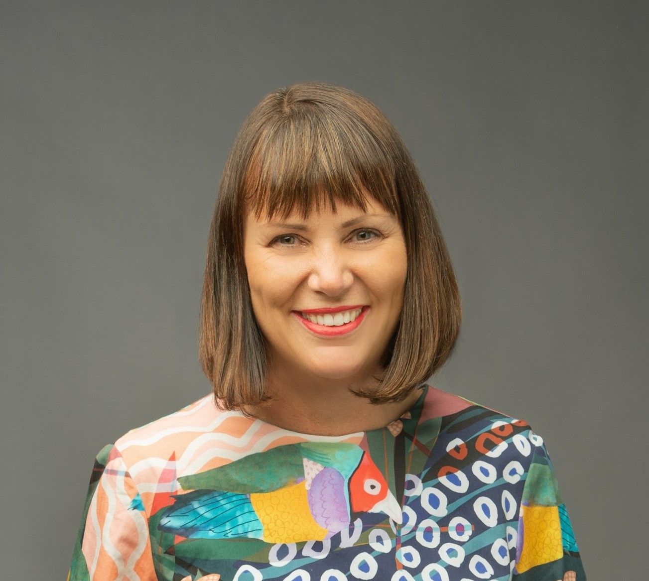 Woman with short brown hair and colourful blouse standing front on and smiling and the camera