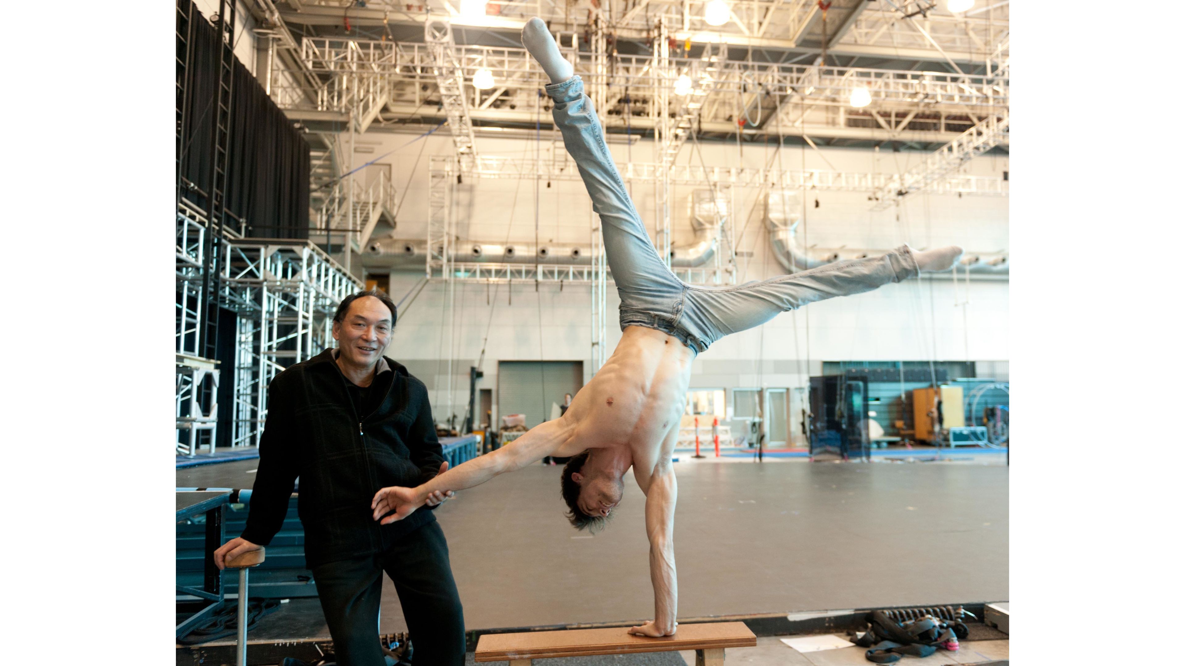 An instructor smiles at the camera while training an acrobat who is performing a one-arm handstand