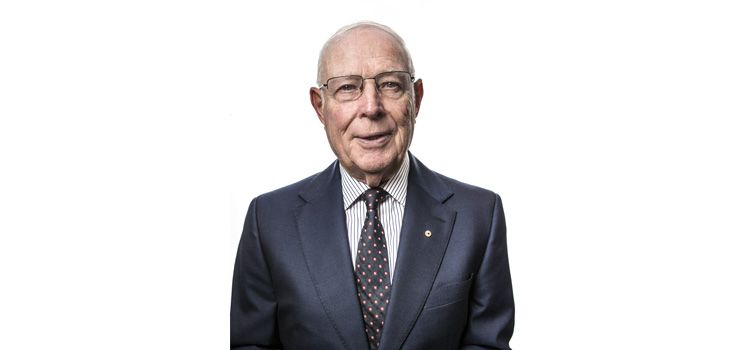 An older man dressed in a suit smiles at the camera in front of a white background
