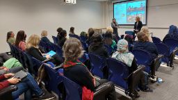 A lecture theatre with women attending a workshop. Two presenters stand in front of the group talking.