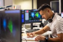Aviation student at a desk looking at a notepad,with flight software on the computers in front of him.