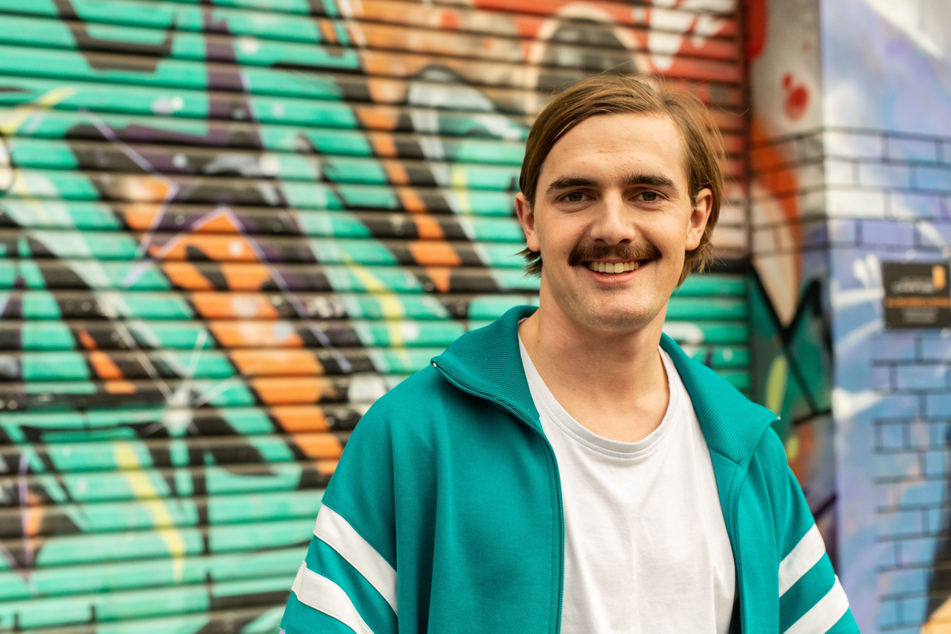 Man with stache standing in front of a graffiti-ed wall, looking pleased with his life choices.