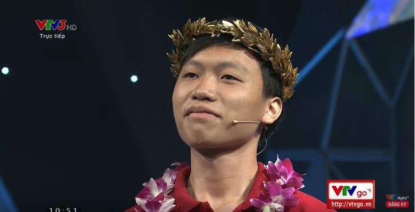 High school student wearing crown of gold leaves, flower chain around his neck and microphone/earpiece. TV logos and time stamp watermarked on shot.