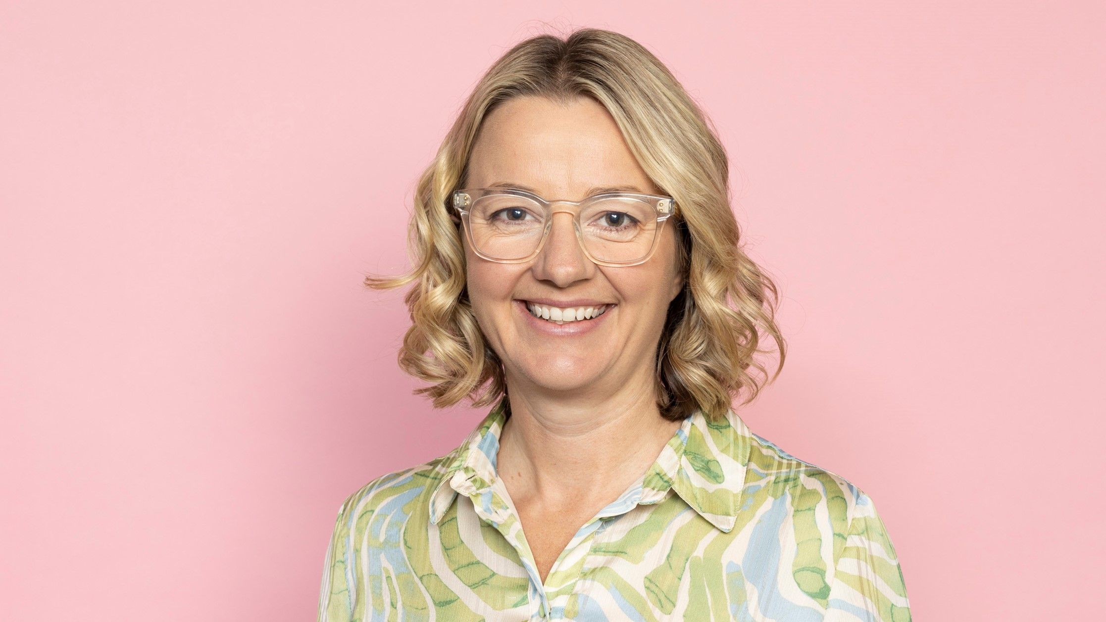 Jen Sharpe wears a pastel green and blue printed dress and clear-framed glasses. She stands smiling in front of a pale pink backdrop
