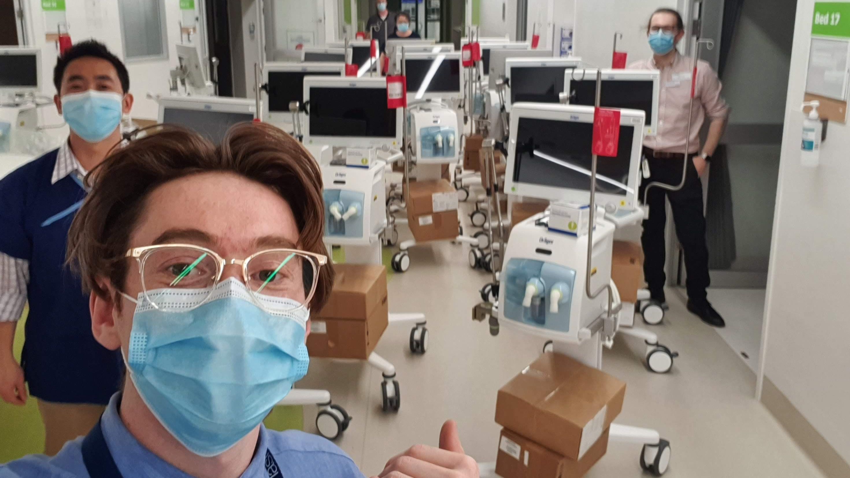 Josh Castle taking a selfie with his thumbs up in a hospital setting. an array of monitors on wheels and two classmates are visible in the background