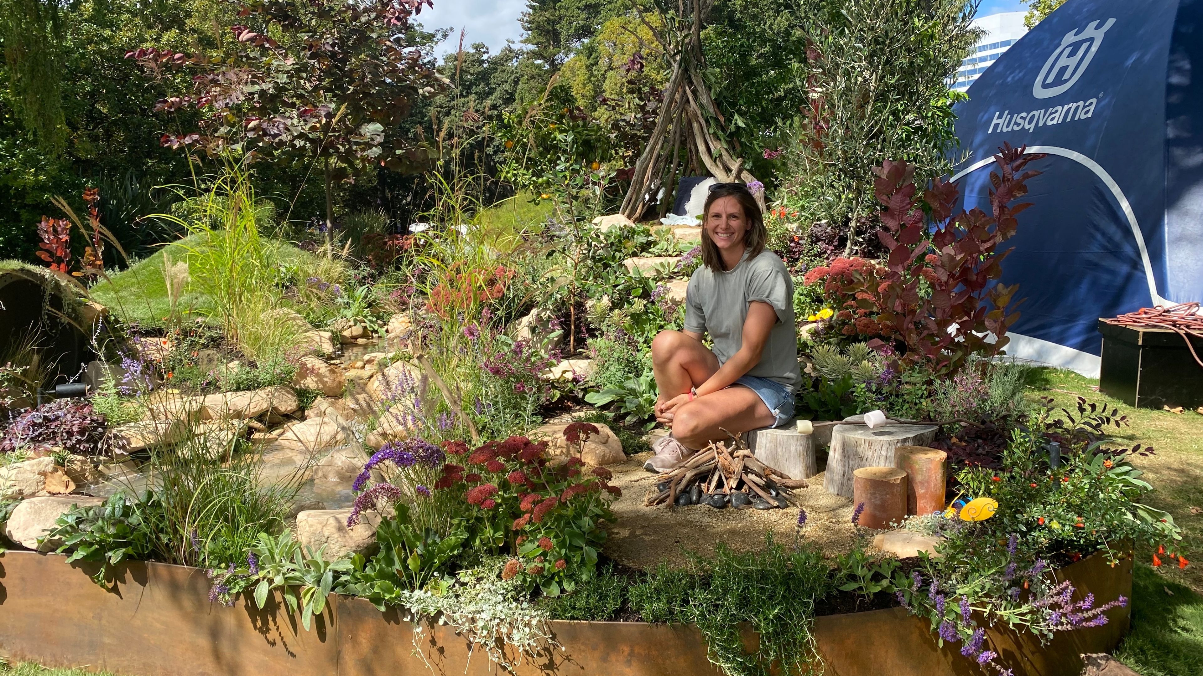 Katie Johnstone sits next to a little teepee fireplace in her emergent garden. She is surrounded by kid sized hills, plants and other fun things to explore