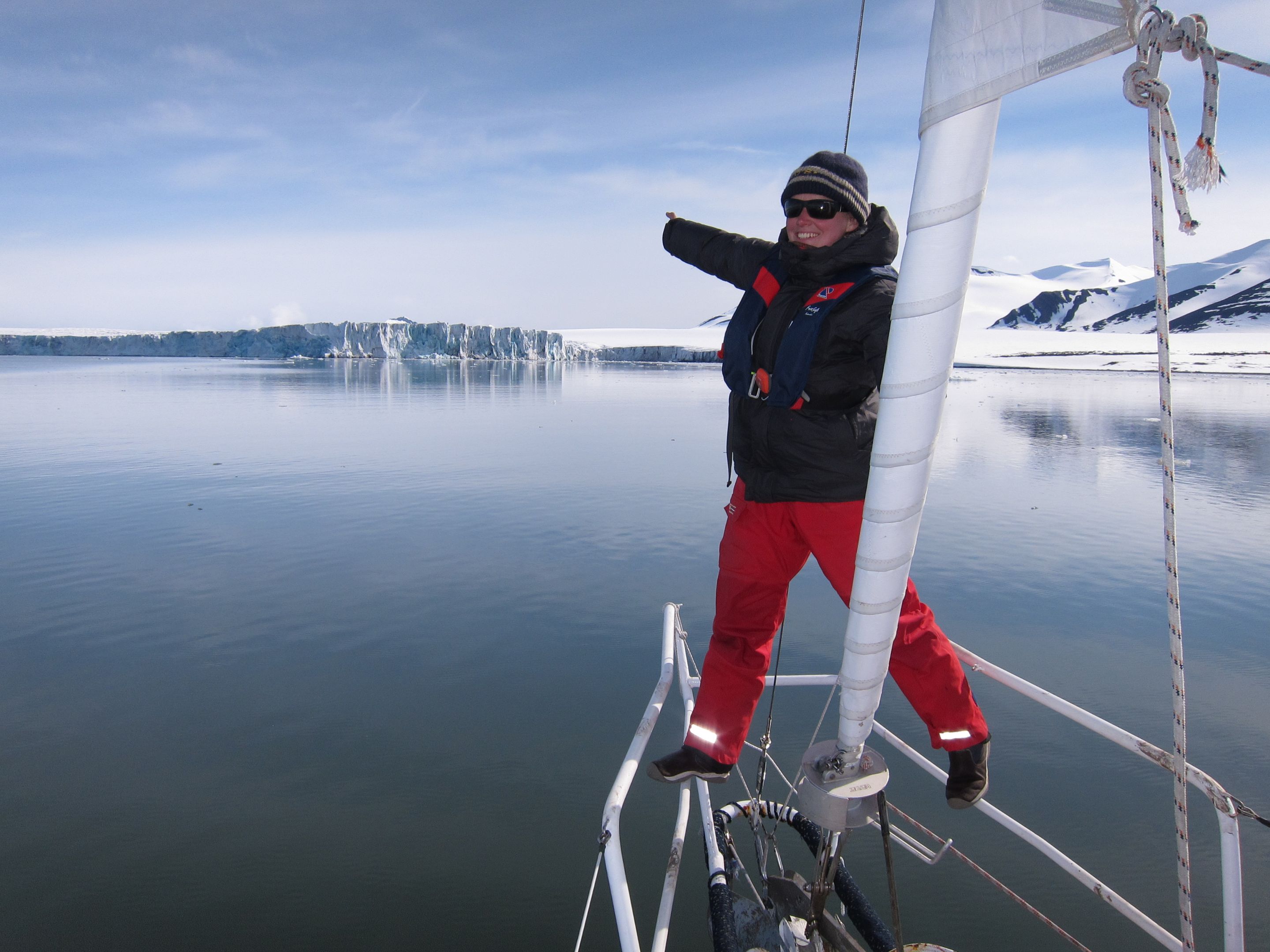 Dr Leonie Suter sailing in Svalbard, one of the world’s northernmost inhabited areas. She stands at the bow of the yacht, pointing out towards an iceberg in the water 