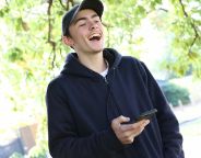 Image of a young man in a black hoodie, black cap, holding a phone, smiling. Image is on a slight tilt.