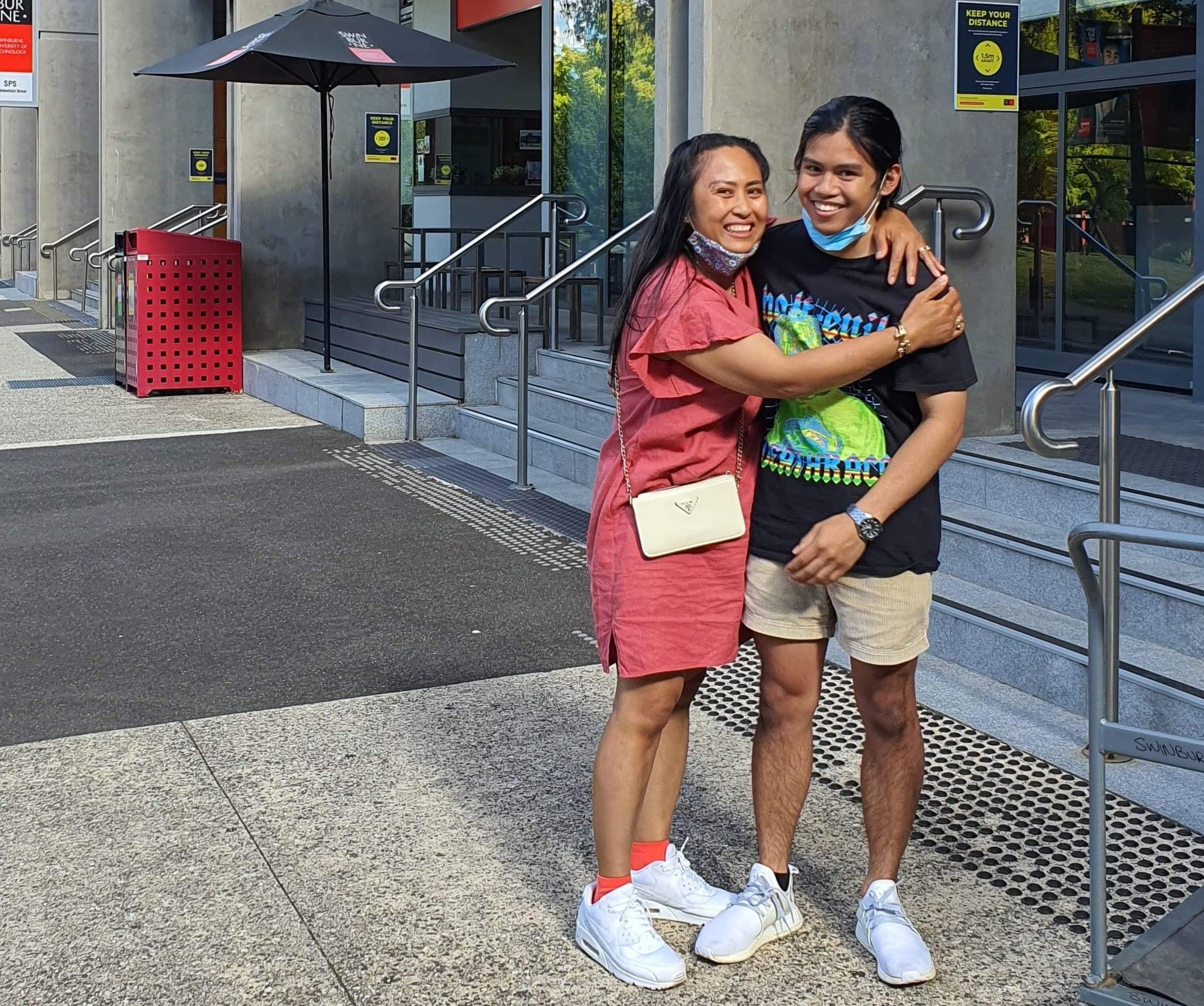 Swinburne student Stephen wears summer clothing and stands outside the concrete steps and glass doors to Swinburne student residences with his mum. His mum is hugging him and they both are smiling happily at the camera. 