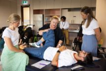 A student lying on a table being shown by his teacher how to assist someone stretch in a classroom with other students.