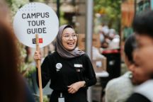 Student holding up a white circular sign on a stick which reads campus tour guide 