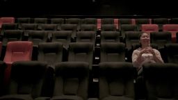 Female student sits in an empty movie theatre, wearing a pink shirt and smiling animatedly.