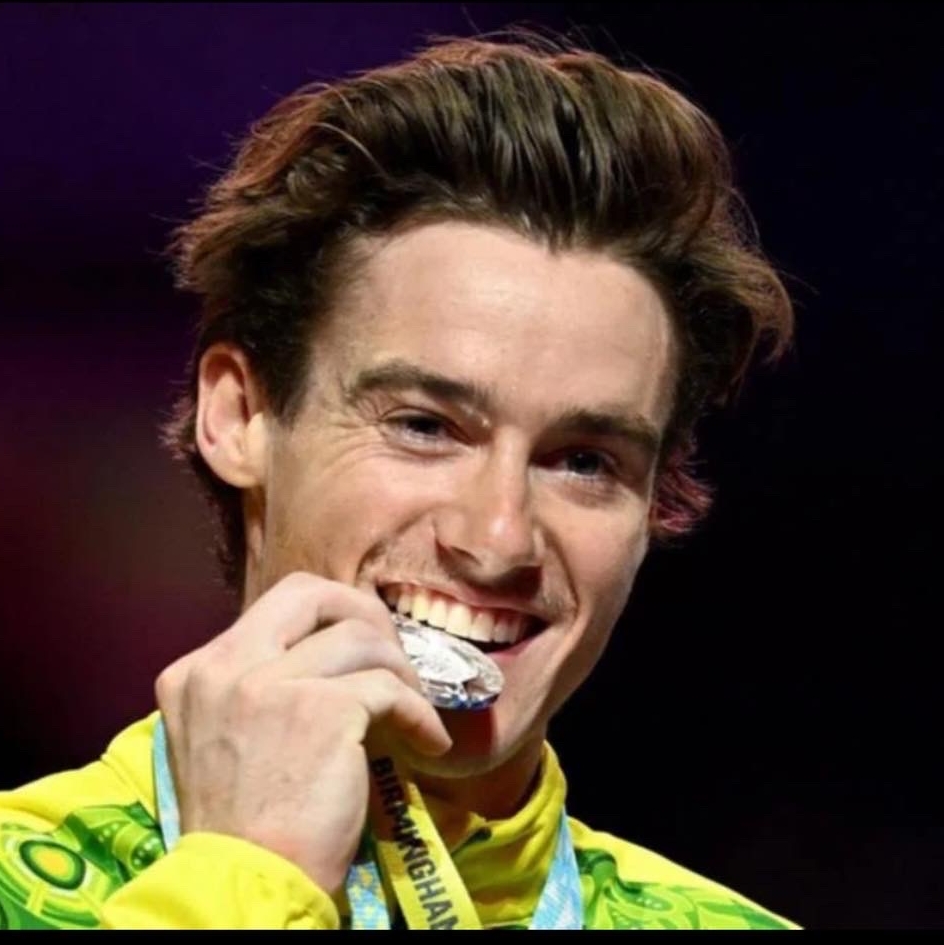 A young man in a green and gold tracksuit is smiling holding a silver medal