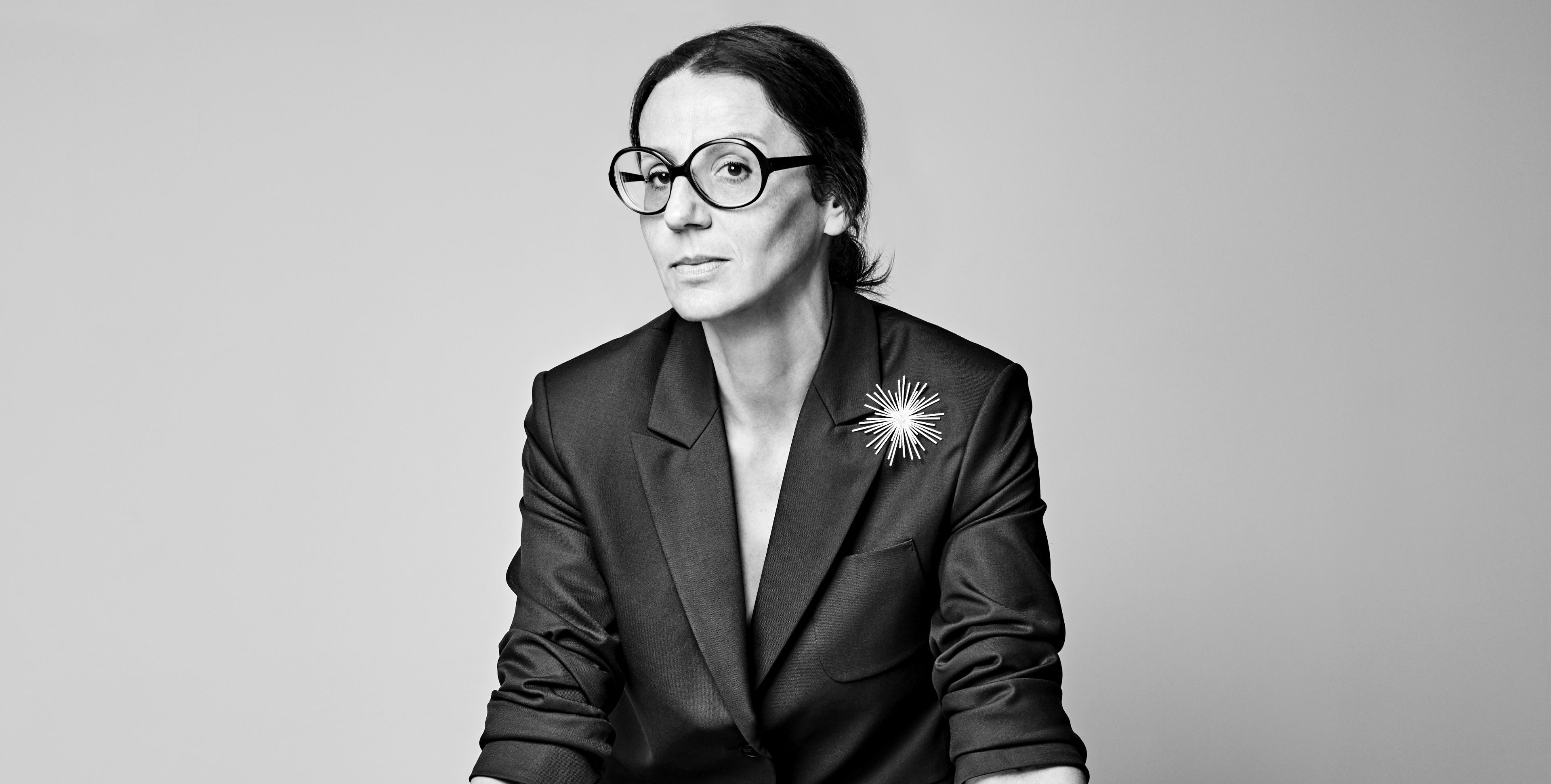 A black and white portrait of Visnja. She wears a simple, clean lined black suit with one of her brooches attached. She is also wearing large, black-framed glasses. She has dark hair and fine features. 
