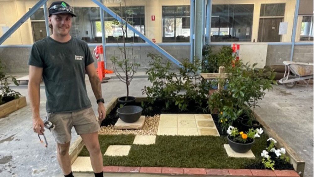 Student stands next to square plot with grass, plants, pavers and flowers
