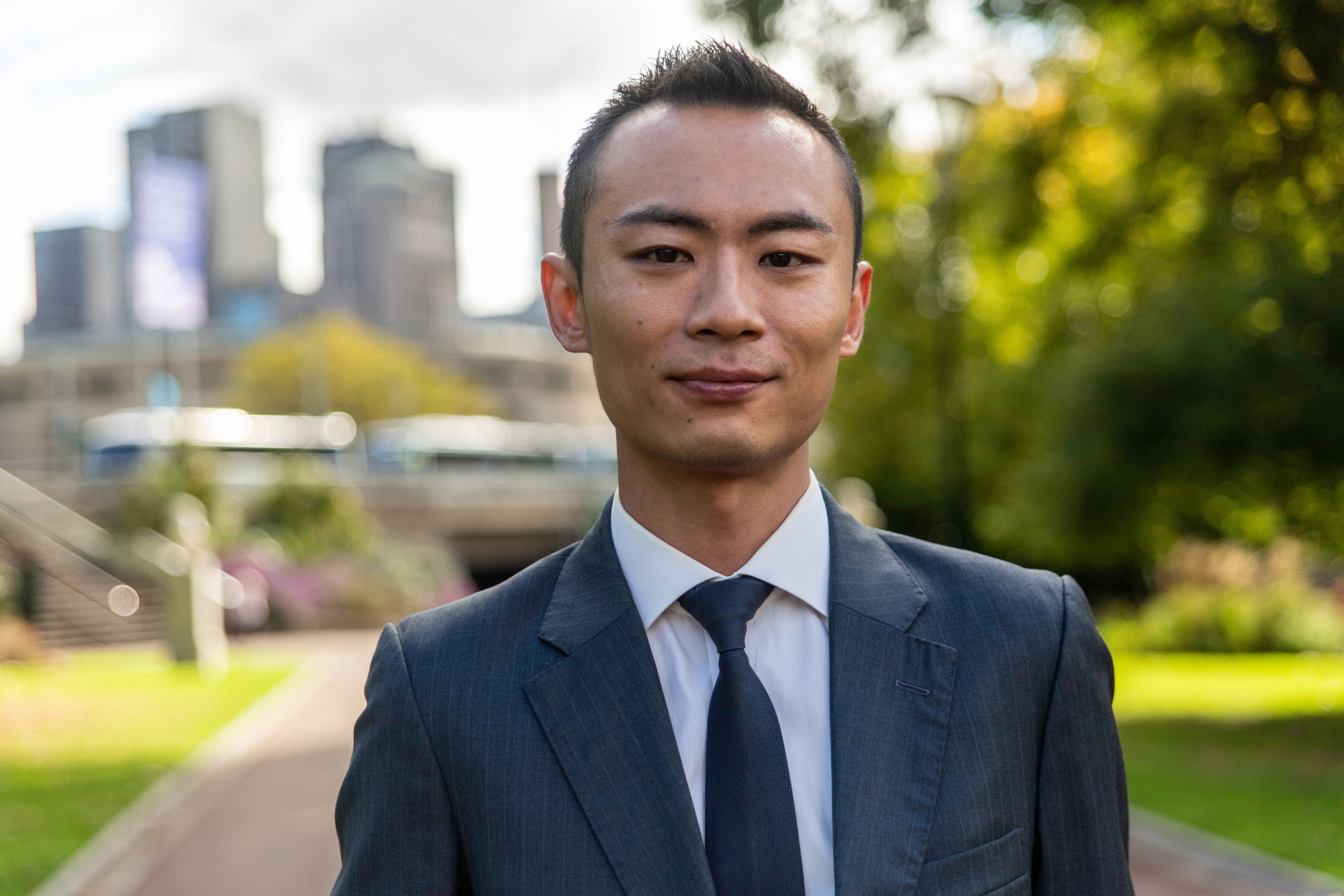 Close up of Christian Tang in suit attire with park greenery and Melbourne CBD blurred in background