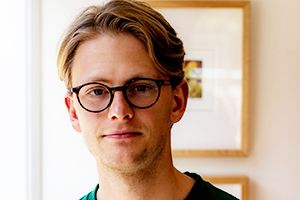 A man with light brown hair and black glasses looks to camera