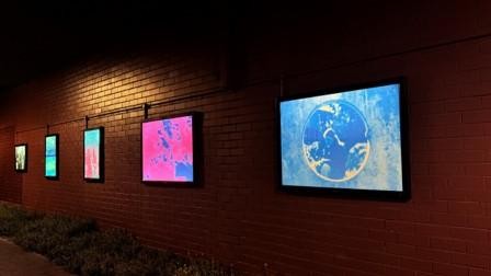 Outdoor installation of illuminated digital artworks mounted along a brick wall.