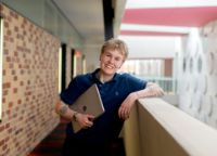 Person standing leaning with their arm on the wall with laptop in hand smiling at camera