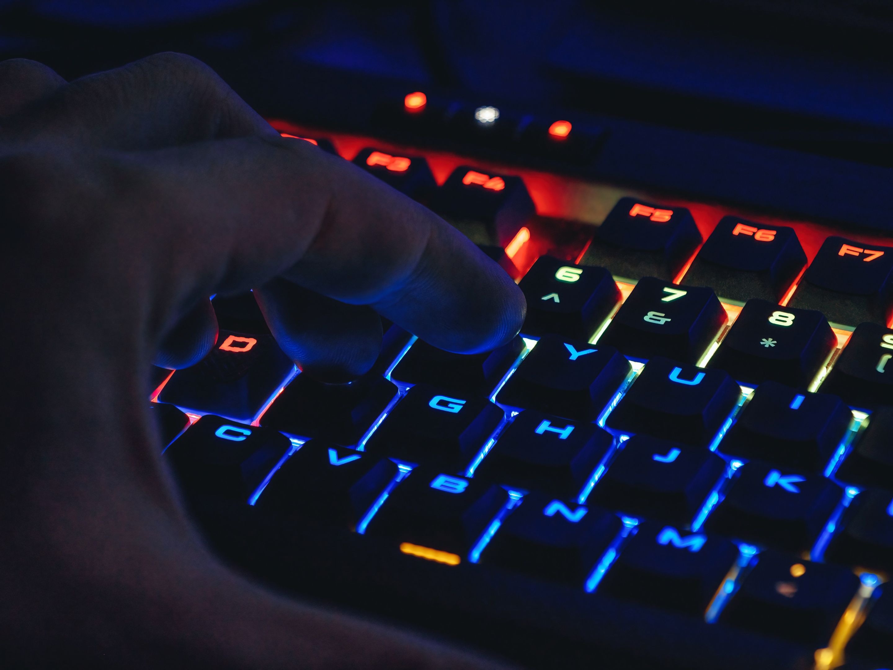 A neon-lit keyboard shine blue, orange and red. A man's hand is poised above it, ready to type. 