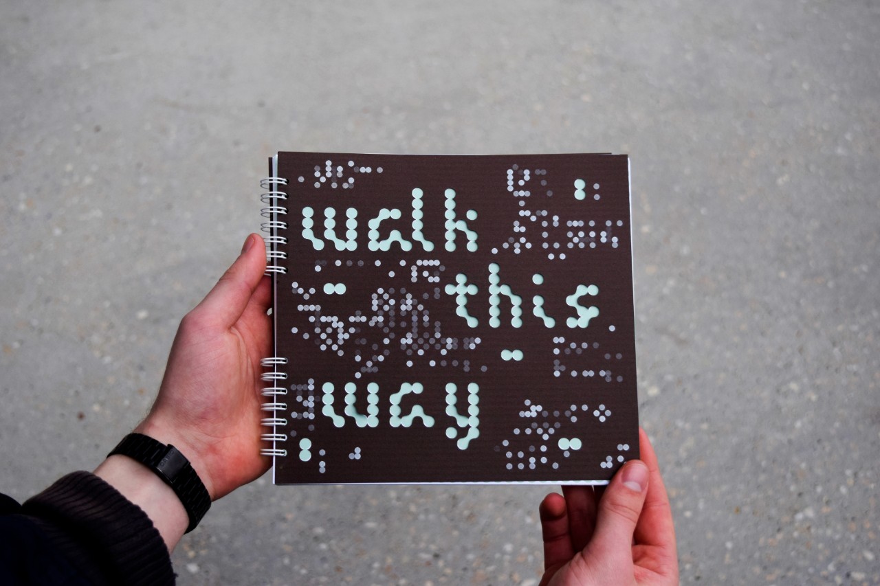 Hands hold a ring-bound publication with a black cover filled with code-like dots. The largest dots are cut out to spell "walk this way"