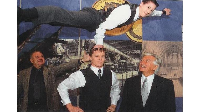 Two men watch on as an acrobat hand balances on another acrobat's head