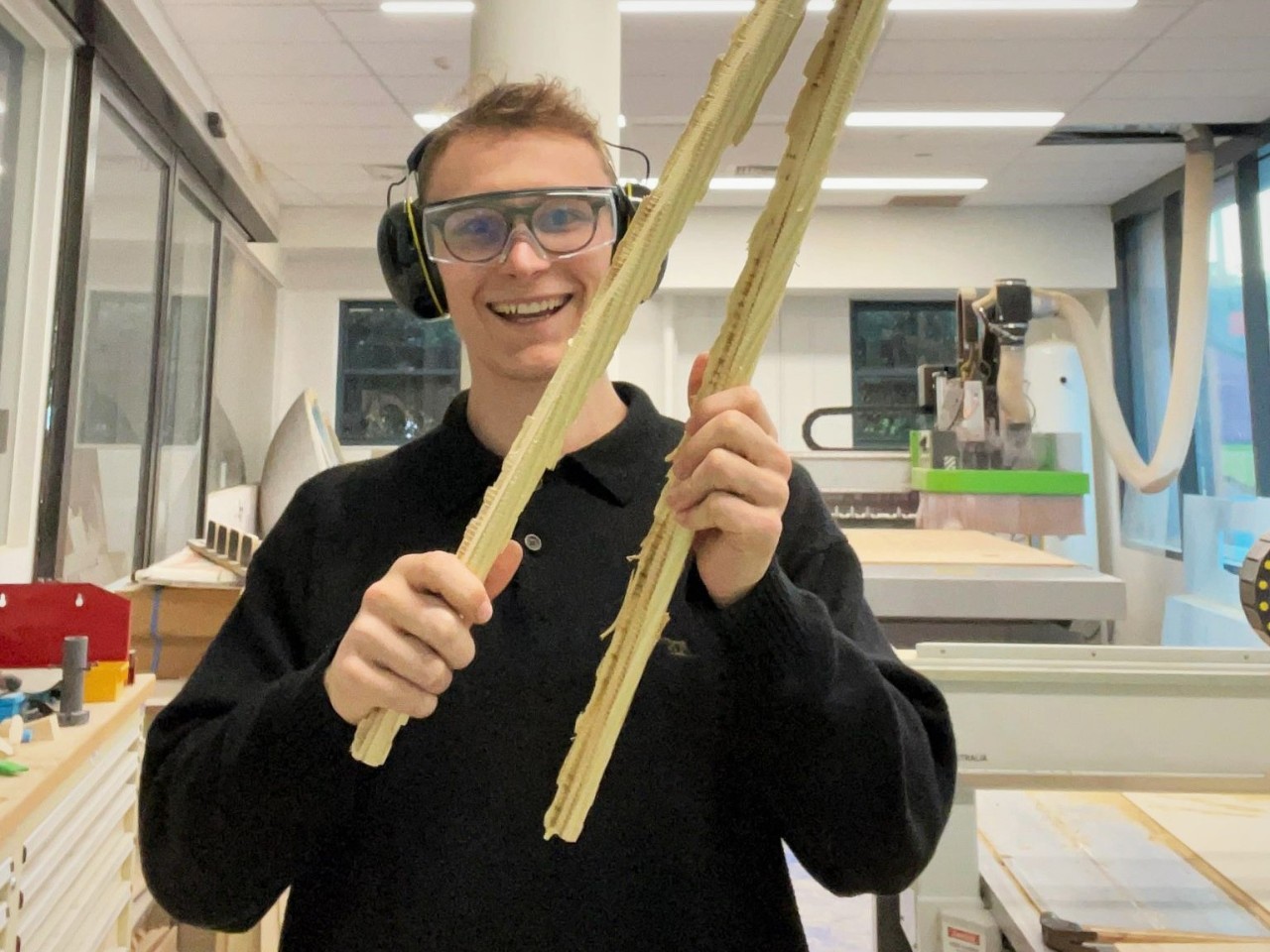A student in safety glasses and earmuffs hold up roughly cut two pronged handlebars 