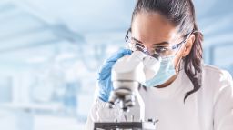 Female medical lab technician in protective glasses, gloves and face mask sits next to a microscope in laboratory.