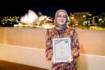 Photograph on a balcony at night of a person in a floral dress holding a framed award with the lights of Sydney and the shape of the Opera House in the background