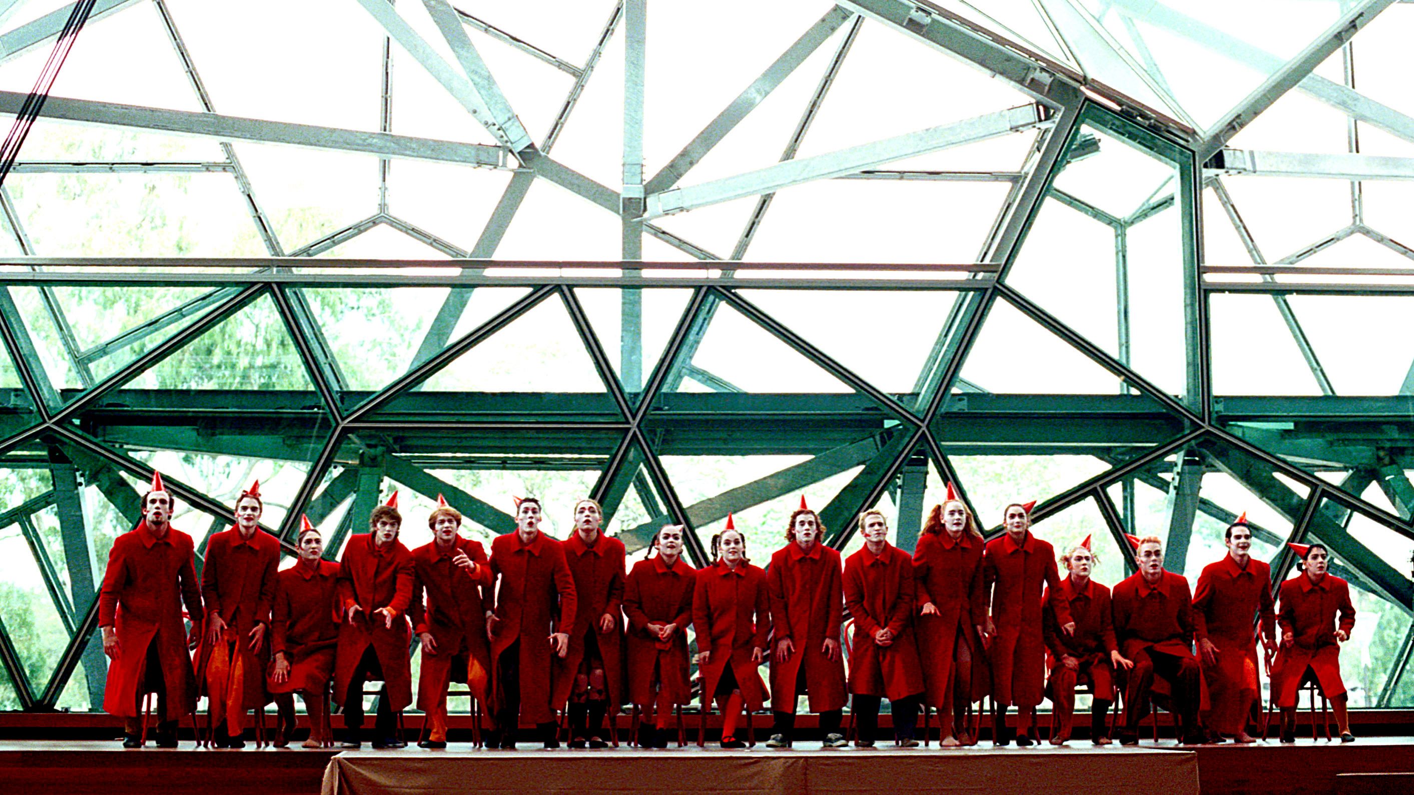 Performers in red costumes pose in front of a large glass window which includes metal rods forming geometric shapes